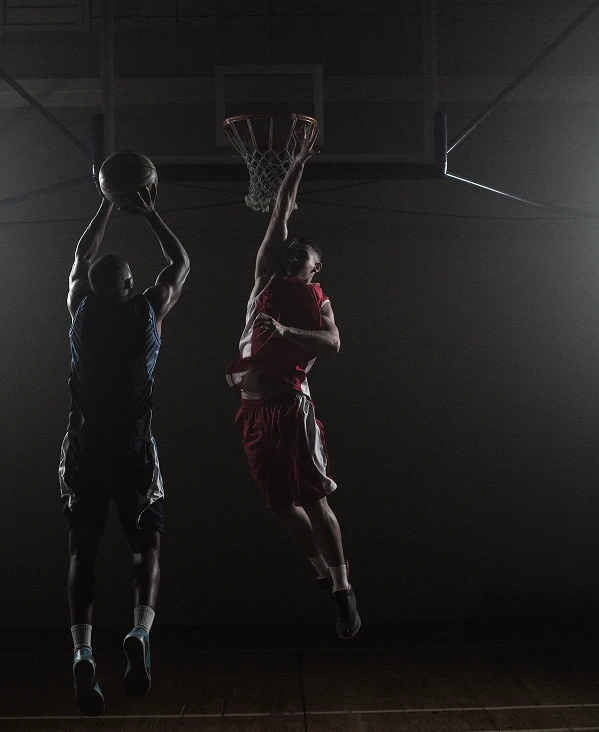 Two basketball players jump near a hoop in a dimly lit gym. One in red attempts a basket, while the other in black tries to block. The scene is intense and dramatic.