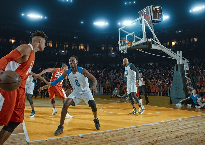 Match de basket en cours ; le joueur en rouge dribble devant le défenseur en blanc. Terrain municipal, concentration intense. Public et lumières vives en arrière-plan.